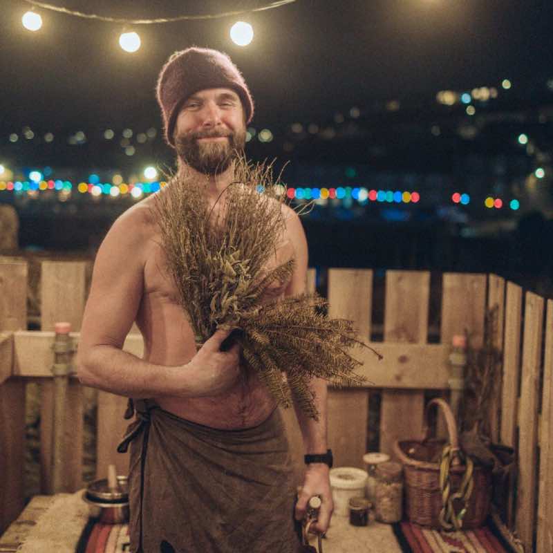 Sauna Master Nick T is standing outside a sauna during a sauna ritual. He is holding a sauna whisk and wearing a sauna hat
