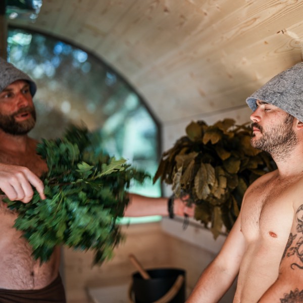 Sauna master Nick T is guiding a sauna ritual. The male participant is wearing a sauna hat and receiving the healing benefits of sauna whisks
