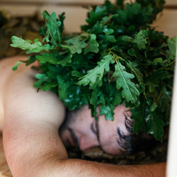 A man is experiencing the nourishing benefits of a sauna whisk treatment. He is laying down with the whisks applied as a hot compress on his head