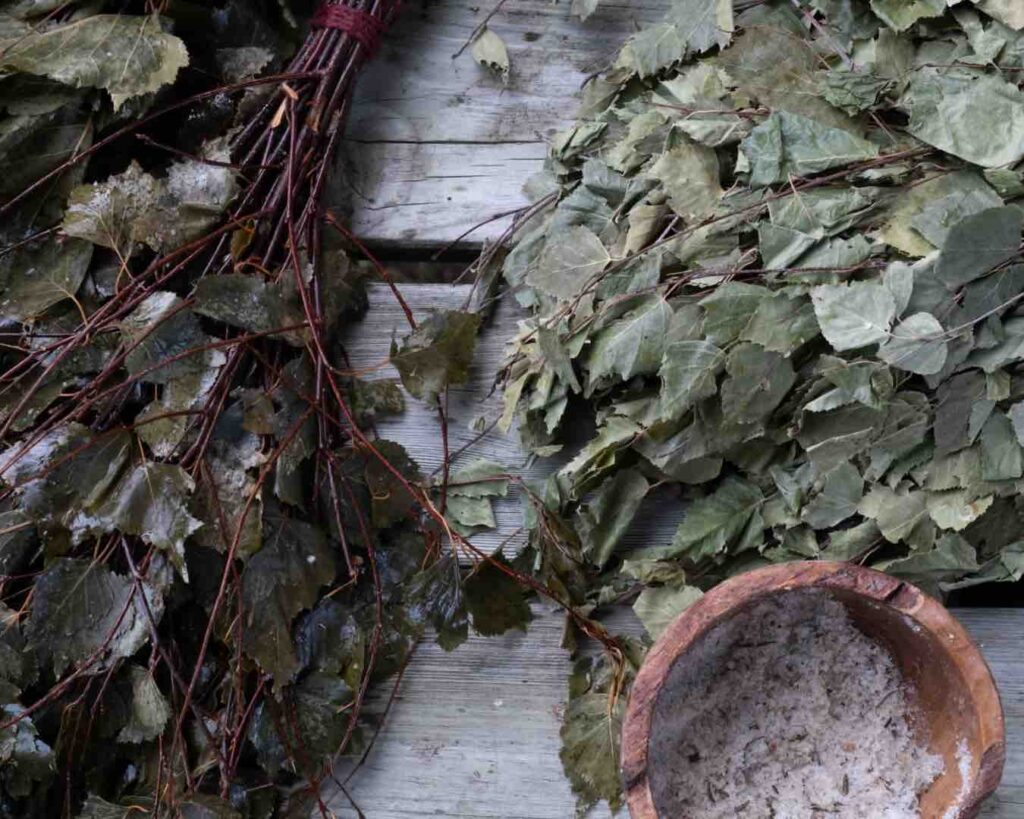 Two sauna whisks made from birch twigs and leaves, they are laying next to a wooden bowl full of a salt body scrub made for the sauna