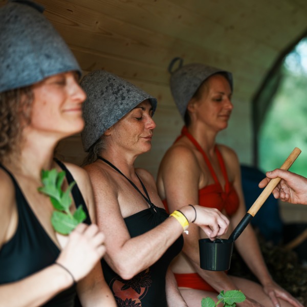 A group of women are enjoying a sauna ritual. They are holding leaves and wearing sauna hats. One is making a sauna wish and setting intentions with a sauna ladle