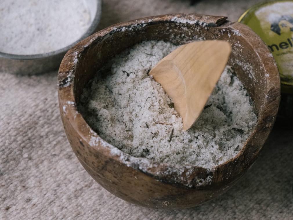 A rustic wooden bowl containing a homemade salt and herb body scrub for use in the sauna and used as part of a sauna ritual. The scrub contains sea salt, rosemary and essential oils and the scoop is hand carved wooden spoon