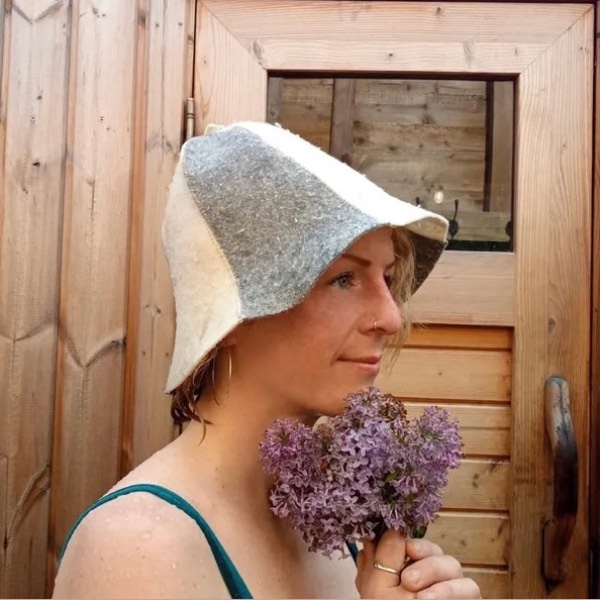 Charlotte Bradman is holding a whisk of Lilac flowers and wearing a sauna hat. She stands outside the sauna