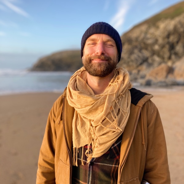 Nick is standing on the beach in Cornwall. The sea is in the background, the sun is shining and the sky is blue