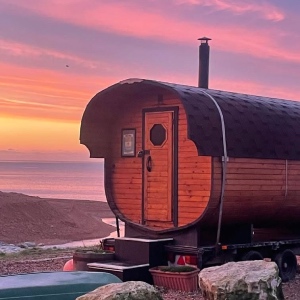A wood-fired mobile sauna sits at the top of the beach in the sunset