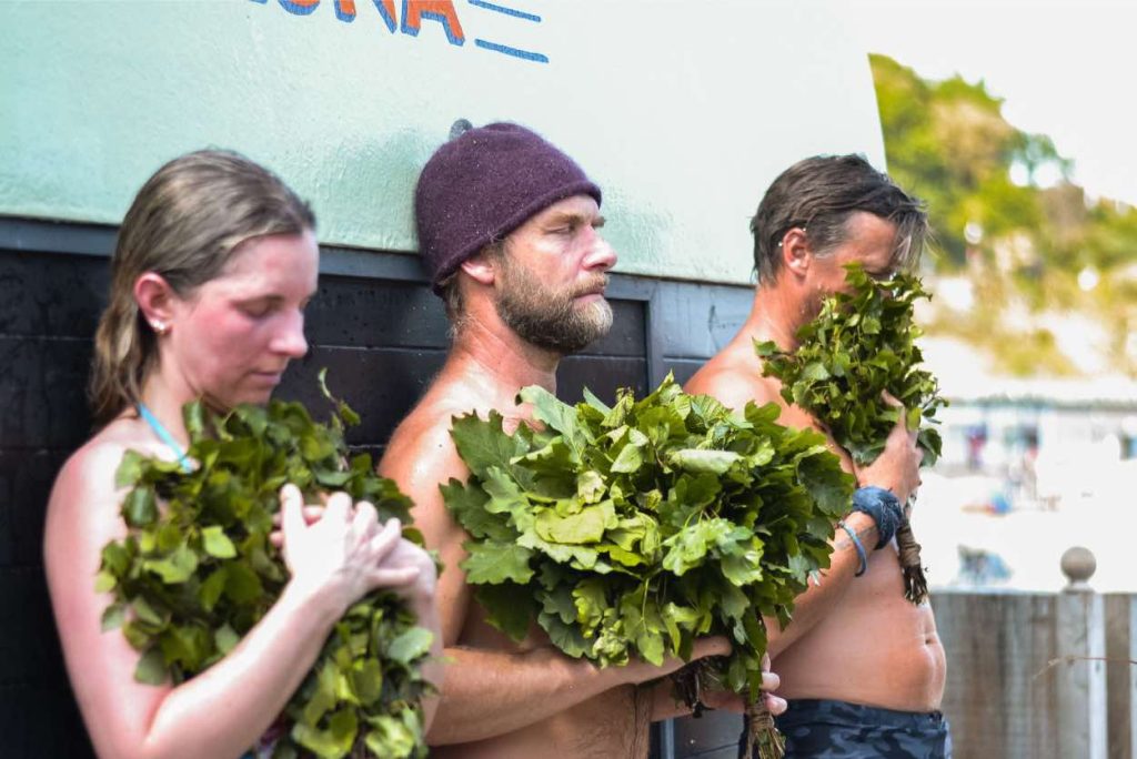 NIck T, sauna master. is guiding a couple in a ritual. They are standing outside the sauna to cool off, holding sauna whisks, small bundles of leaves and twigs
