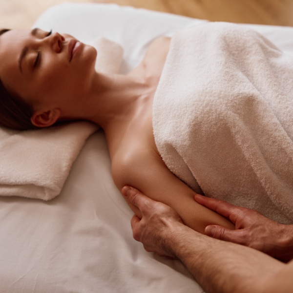 A woman enjoys a relaxing massage treatment from a Massuer. She is laying on a massage couch, wrapped in a white towel