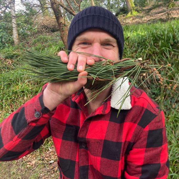 Nick T is foraging in the woods of Cornwall. He is holding some pine leaves in front of of his mouth. The leaves look like a big smile!